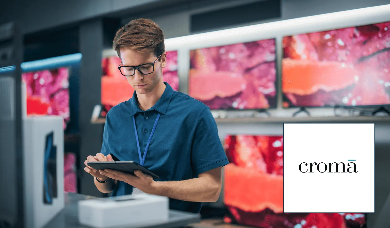 A male electronics store employee with glasses and a blue polo shirt uses a tablet in a store surrounded by televisions. The croma logo is in the bottom right corner.