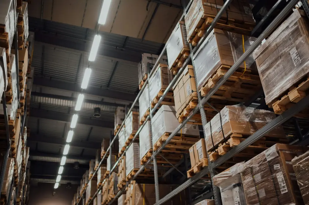 A large, modern warehouse with tall shelves filled with stacked boxes and pallets. The ceiling has rows of bright fluorescent lights.